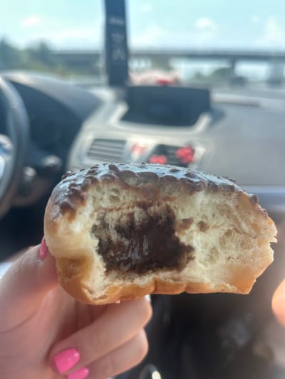 A close-up, eye-level shot shows a hand holding a bitten donut with a chocolate filling. The donut is topped with a chocolate glaze and sprinkles. The background, out of focus, suggests an indoor setting, possibly a car interior, with a glimpse of a road and sky through a window. The location is identified as Tsalapitsa, Bulgaria.