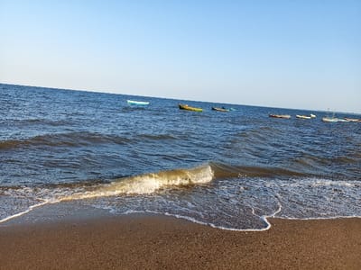 The image depicts a sandy beach with calm, blue water and several small boats anchored in the distance. The image is captured from the shoreline looking out towards the ocean. The location is Praia da Costa do Sol, Maputo, Mozambique, at the coordinates -25.954, 32.611.  There is a small, foamy wave breaking on the beach. The sky is clear and blue. There are no people visible in the image.