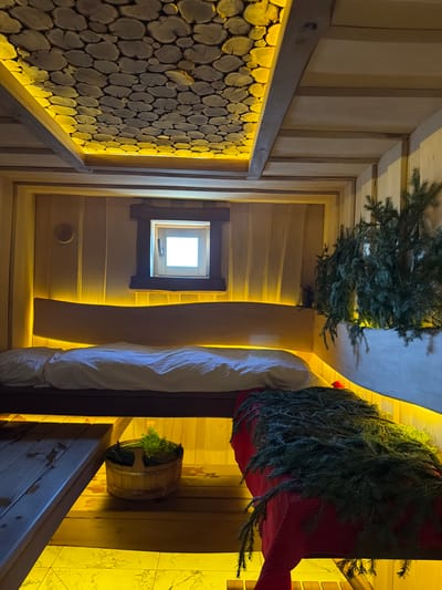 This image shows the interior of a room with wooden walls, likely taken from a lower perspective looking upwards. The location may be in Марусино, Russia. The room appears to be decorated, possibly for a holiday. There are two bunches of evergreen branches, one near the top of the frame on the left and another larger bunch draped over what looks like a bed on the right. A small window is visible in the middle, and a circular pattern of cut wood fills a section of the wall on the left. There is also a small wooden bucket filled with greenery on the right. The room is illuminated with warm, yellowish lighting.