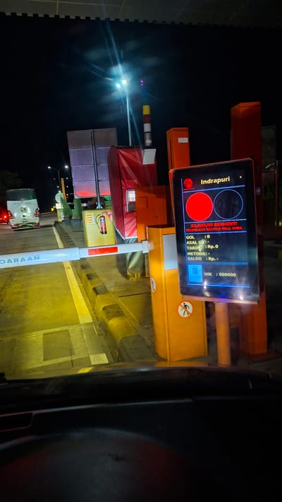 The image captures a nighttime scene at a toll booth, specifically identified as being in Sihomlhok, Indonesia. The foreground shows the interior of a vehicle, looking out through the windshield. A white toll barrier is lowered, blocking the path. To the right of the barrier is an orange toll booth structure with a digital display screen. The screen shows the toll plaza name "Indrapuri" and instructions in Indonesian, indicating to "PLEASE STOP" and "TAP YOUR E-TOLL CARD." It also displays financial information: GOL : 0, ASAL GB : -, TARIF : Rp.0, METODE : -, SALDO : Rp. -, and NIK : 000000. There are also icons of a camera and a person with a prohibition sign on the booth.
In the background, a white bus is stopped at the toll lane. Further back, streetlights illuminate the area, and the night sky is visible. The overall environment suggests a typical highway toll plaza operating at night.