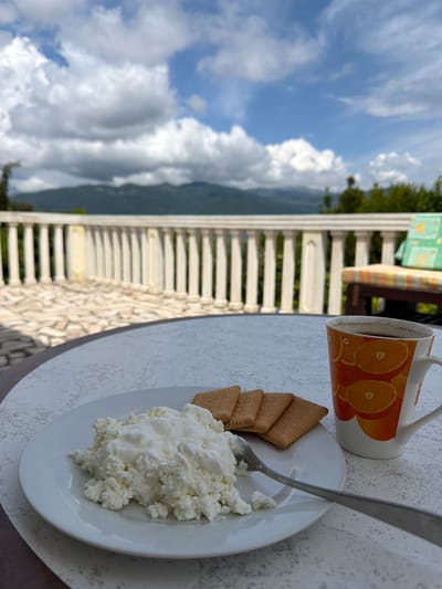 On a table overlooking a partially cloudy sky with mountains in the distance, sits a white plate containing cottage cheese and four rectangular cookies. A silver spoon rests partially in the cottage cheese. A mug with orange slices printed on it stands next to the plate. The location is Krašići, Montenegro. The table is situated on a balcony with white railings.