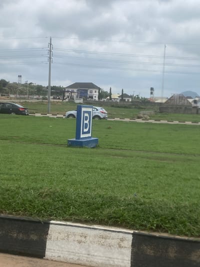 The image presents a wide green field under an overcast sky during daytime in Gosa, Nigeria. In the midground, a prominent blue and white rectangular sign stands on the grass, clearly displaying the capital letter "B". To the right, a curb with distinct black and white stripes borders the grassy expanse.
In the background, several residential buildings and houses are visible, featuring various architectural styles, some with multiple stories. A few cars, including white and dark models, are parked near these structures. Further into the distance, a tall utility pole with numerous power lines stretches across the sky, and another water storage tower can be seen. The scene suggests a developed or developing area with a mix of open green spaces and built infrastructure. No human activity is visible.