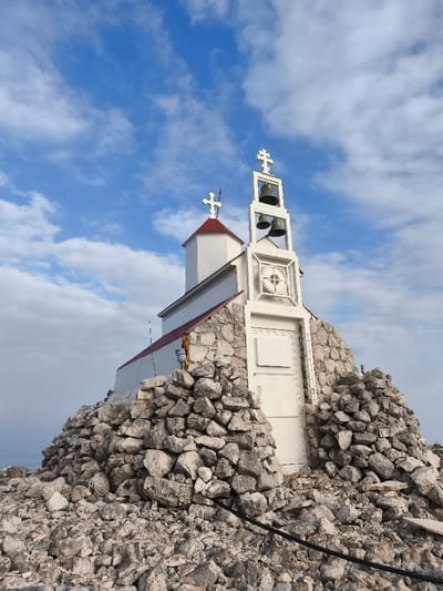 An image taken on a rocky mountain summit in Mali Mikulići, Montenegro, depicts a small, white religious structure under a partially cloudy blue sky. The building features a prominent white facade and a reddish-brown sloped roof on its main section, with a smaller, conical red roof over an adjacent turret or smaller tower topped by a white Orthodox cross. A taller, white, open-frame bell tower stands at the front, containing two dark-colored bells and surmounted by a larger white Orthodox cross. Below the bells, the tower facade incorporates a circular decorative element with a cross design and a rectangular white door at its base. The structure's lower walls are surrounded and partially constructed from a substantial quantity of irregularly shaped, light-colored rocks and rubble, which also covers the immediate foreground. A black electrical cable is visible lying across the rocky ground in the lower right. A small, grey cylindrical object, possibly a sensor or instrument, is attached to the building's side wall near some yellow insulation material. No individuals are present in the scene.