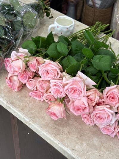 The image shows a bouquet of light pink roses with green leaves arranged on a marble-like surface. Some of the roses are fully bloomed, while others are still in bud form. The stems of the roses are visible, with some still wrapped in clear plastic. To the left of the roses, a white ceramic mug with small red hearts is partially visible. The background is blurred, but appears to be a countertop or table. The lighting suggests it is daytime, and the overall mood is calm and natural. There is no visible text in the image. The location context is Sunzha, Russia, but there are no specific visual cues in the image to confirm this.
