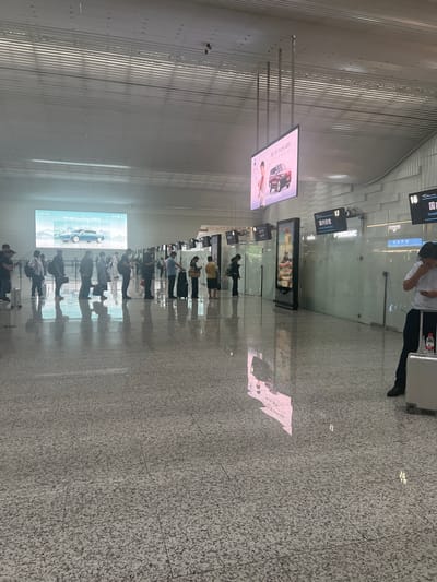 The image shows a busy interior scene, likely an airport terminal, with people queuing in a line. A large screen displaying an advertisement for a car is visible in the background. Several signs with Chinese characters are also present, indicating different sections or services, such as "Domestic Security Check." The floor is polished, reflecting the overhead lights and the movement of people. The architecture appears modern, with a high ceiling and metallic structural elements. The presence of luggage suggests travel. Based on the visible signage with Chinese characters and the overall appearance of a major transportation hub, the location is likely Guangzhou City, China.