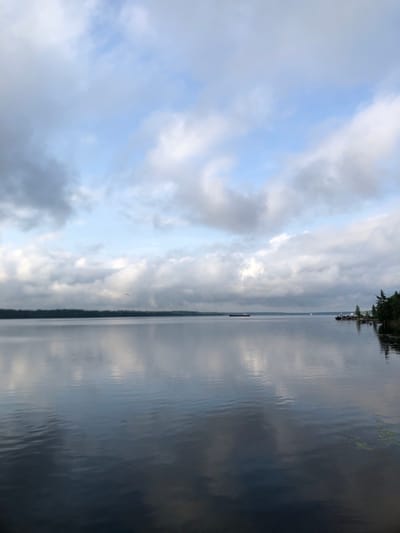 The image shows a body of water, likely a river or lake, in Svirstroy, Russia. The water is calm with gentle ripples reflecting the sky. On the right side of the image, the water surface is dark and reflective, showing streaks of light that suggest either clouds or the sun breaking through them. On the left side, a cloudy sky is visible with patches of blue sky peeking through. The clouds are varied, with some appearing white and fluffy and others darker and more dramatic.
Along the far edge of the water on the right, a dark, tree-lined shoreline is visible, with a few structures, possibly buildings or docks, interspersed among the trees. The overall impression is of a serene and somewhat overcast day, possibly early morning or late afternoon due to the diffused light. The scene evokes a sense of tranquility and natural beauty. There is no visible text in the image.
