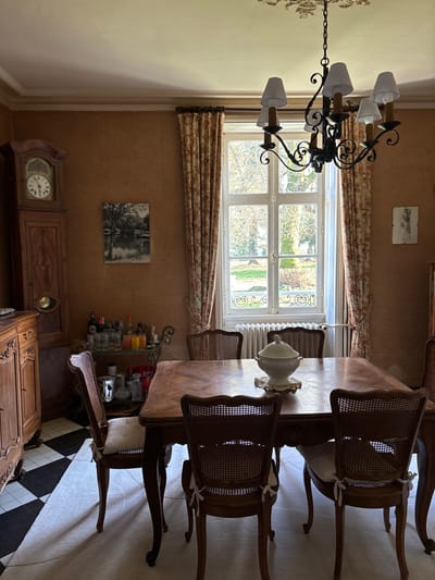 This image presents a classic and elegantly furnished dining room or formal living area, illuminated by natural light from a large window. In the center, a rectangular wooden dining table, featuring a carved base, is surrounded by several traditional wooden chairs. These chairs have cane backs and seats, with some adorned with small white tie-on cushions. A white ceramic tureen, resting on a decorative stand, acts as a centerpiece on the table.

Above, a prominent black wrought iron chandelier, equipped with six shaded lights, hangs from the ceiling, contributing to the room's traditional aesthetic. The warm, textured brown walls display two framed artworks; one is a monochrome landscape, and the other a smaller, lighter piece. Floral patterned curtains flank the multi-paned window, offering a view of a bright outdoor setting with trees and a green lawn, indicative of a garden or estate. A white radiator is visible beneath the window.

To one side, a tall wooden grandfather clock, displaying Roman numerals and showing approximately 8:50 or 9:50, stands next to a wooden cabinet. Further into the foreground, another ornate wooden cabinet or chest with carved details is seen, alongside a bar cart or shelving unit stocked with various bottles and glasses. The flooring in this area features a distinct black and white checkered tile pattern.

No individuals are present, and the scene depicts a static interior. The bright light through the window suggests it is daytime. The decor and furnishings evoke a sense of traditional European elegance, fitting for a home in Étréchy, France.