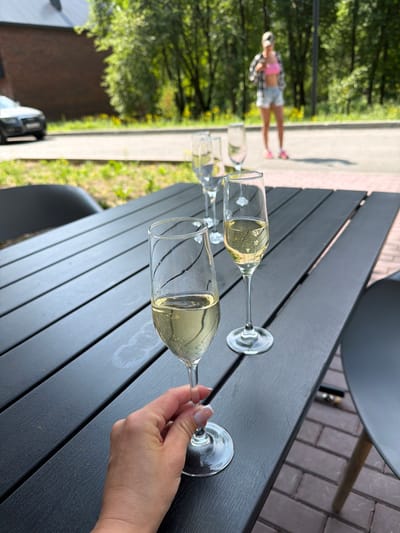 In Morozovo, Russia, a hand holds a champagne flute filled with bubbly champagne, resting on a dark, slatted outdoor table. The table is set with three other empty champagne flutes and another filled flute, suggesting a gathering or celebration. In the background, a blurred woman in a pink top and denim shorts stands among trees, with a car visible on a paved road. The environment appears to be an outdoor seating area, possibly a cafe or restaurant, with a brick-paved ground and greenery. The overall scene conveys a sense of leisure and enjoyment on a sunny day.