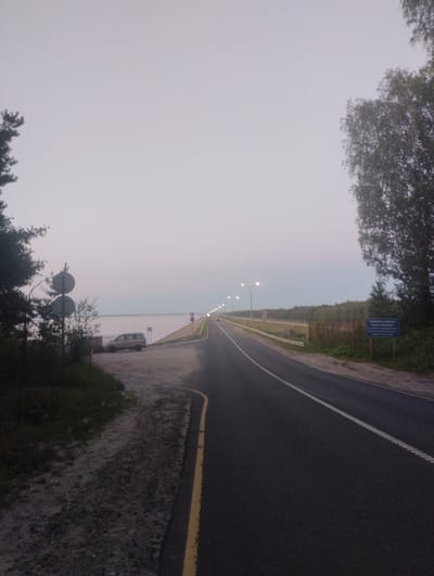 The image captures a view of a road leading to a body of water, likely a reservoir, in Zavolzhye, Russia, during twilight or early morning. On the left side of the road, a silver SUV is parked near a fence and a pole with two circular signs. A dense line of trees borders the right side of the road, which stretches into the distance, illuminated by streetlights. A blue sign with white text is visible on the right, near the trees. The sky is overcast and light, suggesting diffused natural light. The road has white lane markings and a yellow edge line. The overall scene depicts a serene, somewhat desolate, rural roadside environment.