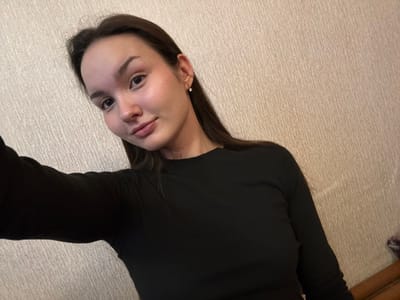 A self-portrait captures a female individual indoors in the city of Bayronovka, Russia. The subject, appearing to be a young adult, has a fair complexion, dark brown hair, and is wearing a black long-sleeved top. A small, clear stud earring is visible in her right ear. She displays a slight, closed-lip smile and maintains direct eye contact with the camera. The background features a light beige wall with a subtle vertical textured pattern. An indistinct dark object, possibly a piece of furniture, is partially visible in the lower right corner.