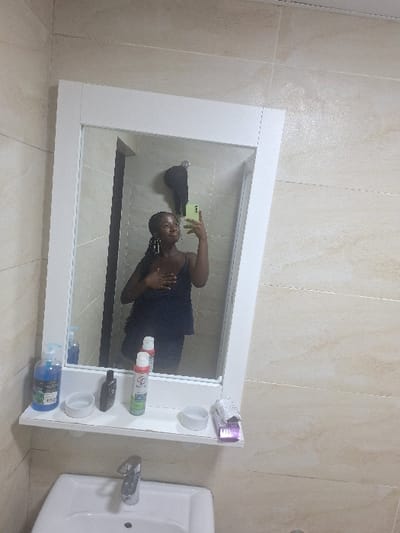 The image displays a modern bathroom interior. The precise location within Close, Nigeria, is not discernible from the visual evidence. A female individual, appearing to be of dark complexion with braided hair, is visible in the reflection of a large, white-framed mirror. She is wearing a dark, possibly navy, tank top and is actively taking a selfie using a light green smartphone held in her raised right hand. Her left hand rests on her chest, and she is smiling.
The mirror features an integrated white shelf below the reflective surface. On this shelf, from left to right, are: a blue pump bottle of liquid, a black cylindrical bottle, a green and white aerosol can, two small white circular dishes or containers, and a small purple object wrapped in white material. Below the shelf and mirror is a white rectangular sink with a silver-colored faucet. The bathroom walls are covered in light beige or cream-colored tiles with visible horizontal grout lines. Reflected in the upper left portion of the mirror, a dark garment or object is seen hanging on a wall hook.