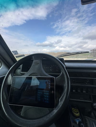 The scene depicts the interior of a vehicle, specifically from the driver's perspective, looking out through a visibly dirty windshield, located in a rural area near Tsenovo, Bulgaria. A black Huawei tablet rests horizontally on the vehicle's black, four-spoked steering wheel, which bears "LADA SPORT" on its central hub. The tablet's screen is reflective, showing distorted reflections. Behind the steering wheel, the vehicle's instrument cluster is partially visible, indicating a maximum speed of 180 km/h. To the right, the dark dashboard features air vents, various control buttons, and a storage compartment containing a yellow tape measure and a small blue object. Two windshield wiper blades are present at the bottom of the windshield. Outside the vehicle, a flat, open landscape is visible under a blue sky with scattered white clouds. A dirt or gravel path extends forward, flanked by low green vegetation. A distant line of bare deciduous trees marks the horizon. No individuals are visible.
