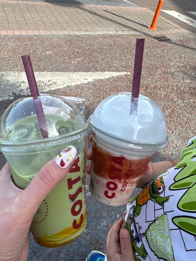 In Carlow town, Ireland, two Costa Coffee drinks are held by individuals. The drink on the left is a green matcha beverage in a clear plastic cup with a dome lid and a purple straw. A hand with decorated fingernails, featuring a ladybug, bee, and flowers, is holding this cup. The cup has red "COSTA" lettering on its side. To the right, a strawberry-flavored drink in a similar cup with a dome lid and a purple straw is visible. This drink has visible layers of strawberry and cream. The background shows a textured ground of small red pebbles and grey paving stones, with a bright orange traffic cone in the upper right corner. A person wearing a green shirt with a Teenage Mutant Ninja Turtle print is partially visible on the right side of the image, with their hand reaching towards the strawberry drink.