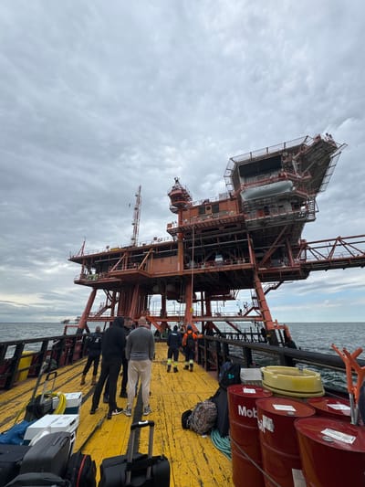 The image depicts an industrial maritime scene on an overcast day, likely off the coast of Marina di Ravenna, Italy. A substantial offshore platform, constructed from reddish-brown metal lattice, dominates the left side of the frame. This multi-level structure features numerous interconnected platforms, pipes, and tanks, including a prominent cylindrical tank on an upper section and a communication mast. The text "REW-C" is visible on a lower portion of the platform.

In the foreground and extending to the right, the bright yellow deck of a ship or support vessel is visible. Several people are standing on the deck; some wear safety vests and hard hats, while others are in casual clothing, mostly with their backs to the viewer. Their presence, along with numerous pieces of luggage such as backpacks, duffel bags, and wheeled suitcases scattered on the deck, suggests a crew change or transfer operation is taking place.

Stacked on the deck are several red industrial barrels, with "Mobil oil" branding clearly visible on some. A yellow, barrel-shaped object or buoy is also present. Other equipment, including ropes and small white containers or toolboxes, is also on the deck. The calm, slightly choppy sea stretches into the background under a cloudy sky, indicating daytime with diffuse lighting.