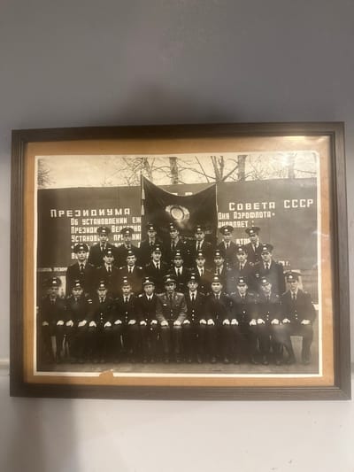The photograph is a black and white group portrait of approximately 30 men, all wearing uniforms and caps. The men are arranged in rows, with some seated and others standing behind them. They appear to be young adults. In the background, there is a large banner with Cyrillic text and a Soviet emblem. The text on the banner includes "СОВЕТА СССР" (Council of the USSR) and possibly other official or organizational inscriptions. The overall scene suggests a formal gathering, likely an official event or graduation ceremony associated with a Soviet-era institution. The photograph is framed with a dark wooden border. The setting appears to be outdoors, with some trees visible in the background. The context provided about Lapės, Lithuania, does not directly appear in the visual information of the photograph itself.