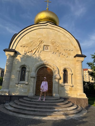 The image shows a woman standing on the steps of a church with a golden dome. The church is made of light-colored stone and has intricate carvings. The woman is wearing a pink outfit and a pink hat. The sky is blue with some clouds. There are trees and buildings in the background. The church appears to be the Church of the Assumption of the Blessed Virgin Mary in Kirishi, Russia.