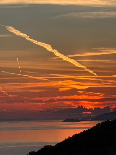 The image depicts a dramatic sunset over the Adriatic Sea, likely viewed from a vantage point in or near Zagora, Montenegro. The sky is ablaze with vibrant orange and red hues, a result of the setting sun interacting with atmospheric conditions. Multiple contrails from aircraft are visible, crisscrossing the sky and catching the light. One particularly prominent contrail stretches diagonally across the upper portion of the frame, appearing illuminated and textured.

Below the fiery sky, the calm sea reflects the warm colors of the sunset, creating a luminous expanse. In the distance, the silhouettes of islands or peninsulas are faintly visible against the horizon. The foreground is dominated by the dark, silhouetted outline of a lush, tree-covered hillside, which creates a stark contrast with the bright sky and sea. No people or specific structures are identifiable in the scene. The overall impression is one of natural beauty and the powerful visual spectacle of a coastal sunset.