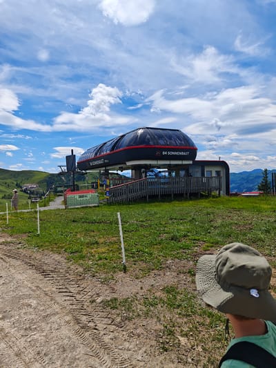 A boy wearing a green bucket hat and a green t-shirt is facing away from the camera, looking at a ski lift station. The ski lift station is black and red with a large glass-enclosed cabin. Text on the cabin reads "B4 SONNENRAST 1776 m". In the background, there are rolling green hills and mountains under a bright blue sky with scattered white clouds. There is a dirt path in the foreground with tire tracks. A woman in a light-colored dress and hat stands to the left of the ski lift station. The scene appears to be at a mountain resort in Kirchberg in Tirol, Austria.