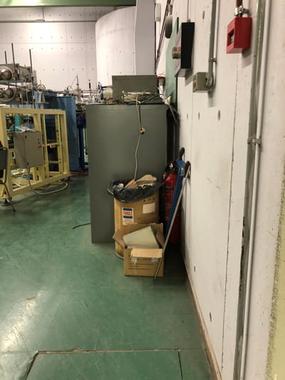 The image captures an indoor industrial or laboratory setting, characterized by utilitarian design and various equipment. The scene is dominated by a large, dark grey cabinet or machine positioned against a light-colored wall. The floor is a distinct light green, possibly an industrial epoxy or painted concrete.
Along the upper part of the wall, several pipes and electrical conduits run horizontally. Mounted on the wall are a red fire alarm pull station and various grey electrical boxes or components. To the right of the large grey cabinet, a red fire extinguisher leans against the wall, partially obscured by several cardboard boxes. One box is clearly marked with the word "FRAGILE" and a "THIS SIDE UP" arrow symbol. Another box or item behind it has handwritten text that appears to be "S. 2019-19". A black trash bag rests on one of the boxes.
Further into the background, other industrial machinery and equipment are visible, including what looks like a yellowish metal rack or frame. The lighting suggests an artificial indoor environment, with no discernible natural light. No people are present, and no specific activity is taking place; the image presents a static view of the space. The overall impression is one of a functional, possibly cluttered, work or storage area in Agia Paraskevi, Greece.