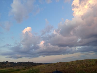 The image depicts a landscape with rolling hills under a dramatic sky filled with clouds. The ground is covered in dry grass and soil, with some patches of green vegetation. In the distance, a range of dark, rocky hills forms the horizon. The sky is a mix of blue and gray, with several large clouds, some of which are tinged with pink and yellow from the setting sun. The overall impression is one of a vast, open natural environment. The location is identified as Баянауыл ауылдық округі, Kazakhstan.