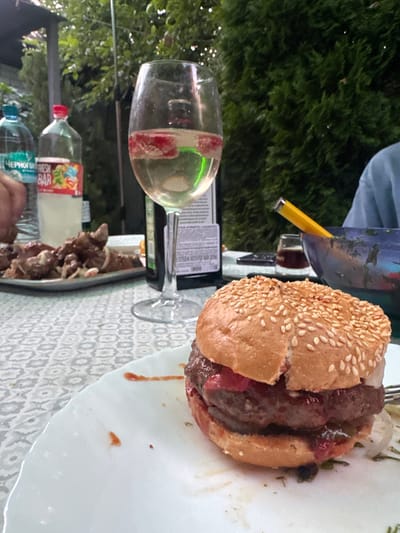 This is a close-up shot of a hamburger on a white plate in an outdoor setting, likely a backyard or patio. The hamburger is the primary subject, with its top bun slightly askew, revealing layers of meat and toppings. The plate has a few red splatters, presumably from ketchup or sauce. In the background, a glass of white wine with fruit slices sits on a table covered with a patterned tablecloth. Several bottles of water and what appears to be a bowl of salad with a yellow utensil are also visible. The environment suggests a casual dining or gathering situation, possibly during a warm weather season, given the greenery and outdoor ambiance. There are no visible people, but their presence is implied by the setup. The location is identified as Pribrezhnyi, Russia.