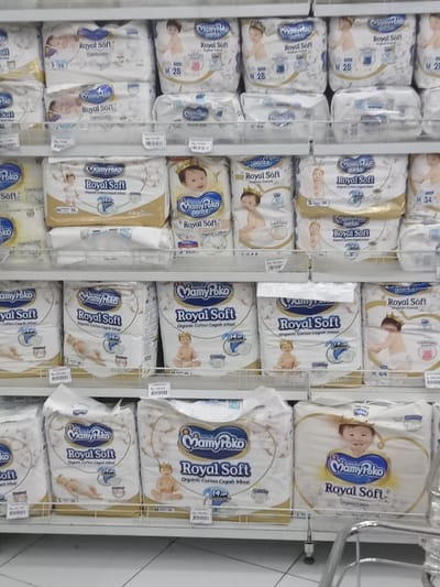 This image depicts an interior view of a retail establishment, likely a supermarket or minimarket, located in Nganjuk, Indonesia. The scene is dominated by multiple white metal shelves, extending from the top to the bottom of the frame, fully stocked with various packages of "MamyPoko Royal Soft" brand baby diapers.
The shelves contain numerous identical packages arranged in rows and stacks. Product packaging is primarily white with blue and gold accents, featuring images of babies, some adorned with small crowns. Text on the packaging, visible in Indonesian, includes "Royal Soft," "Organic Cotton Cergah Iritasi" (Organic Cotton Prevents Irritation), and product sizes such as S 28, M 28, M 34, and 14 pcs. Price tags displaying values in Indonesian Rupiah (Rp), such as "Rp 19.350" and "Rp 179.279," are affixed to the front edge of several shelves beneath the corresponding products.
The environment includes overhead artificial lighting, likely fluorescent, illuminating the display. The floor appears to be constructed of light-colored tiles with visible grout lines. A chrome-plated metal handle, characteristic of a shopping cart, is partially visible in the bottom right corner of the frame. No individuals are present in the scene.