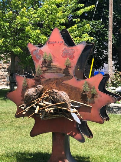 A reddish-brown metal sculpture in the shape of a maple leaf stands on a grassy lawn in Severn, Canada. The sculpture depicts a landscape with miniature trees and a winding path. Two beaver figures are prominently featured, constructed from rough, textured material, and are positioned in front of a pile of sticks, resembling a beaver dam. The sculpture is illuminated by natural daylight, casting shadows on the surrounding grass. The background includes a stone wall, trees with lush green foliage, and a wooden utility pole with wires. A bright yellow object, possibly a flag or marker, is attached to the pole. The overall scene suggests an outdoor public art installation.