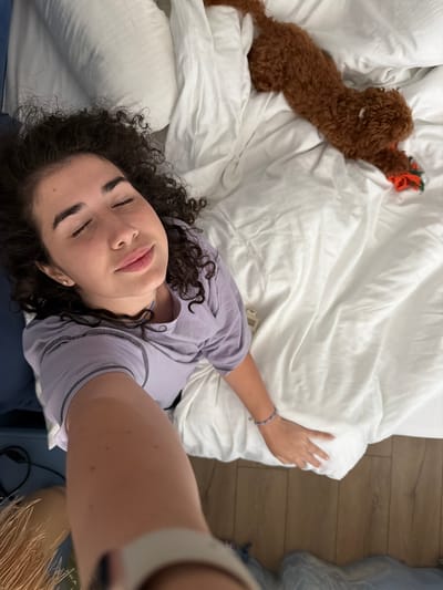 The image shows a woman lying on a white duvet with her eyes closed and a content expression. A small brown dog is lying on the duvet behind her, playing with a toy. The scene appears to be in a bedroom, possibly in the city of Banya, Bulgaria, given the context provided. The woman is wearing a light purple t-shirt. The room has wooden flooring and a blue headboard.