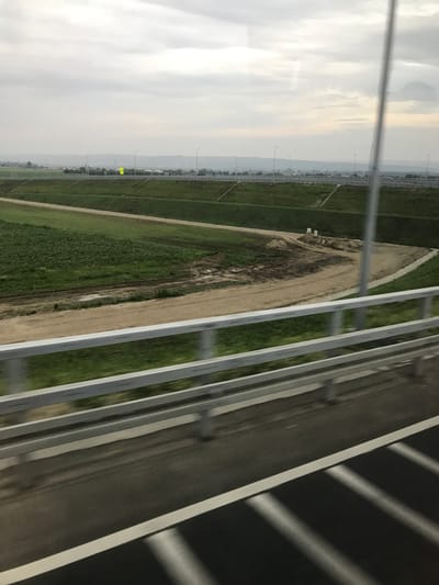 The image depicts an exterior daytime scene captured from a moving vehicle, looking over a highway guardrail towards a landscape within or near Adjud, Romania, under an overcast sky.
In the immediate foreground, the asphalt surface of a road is visible, marked with a solid white line on its right edge. Diagonal light-grey streaks appear on the dark asphalt, indicative of motion blur. Above the road, a silver-colored metallic guardrail with two horizontal rails extends horizontally across the lower third of the frame, also exhibiting slight motion blur. A vertical, silver-colored pole is present on the right side of the image, extending from the bottom edge upwards, partially obscuring the view.
Beyond the guardrail, a sloped embankment covered in green grass descends into a flatter, expansive area. This area comprises sections of dark green vegetation, likely cultivated fields, interspersed with patches of light brown bare earth or dirt. One dirt patch shows distinct tire tracks and small mounds of earth, with two small white rectangular blocks or markers present. Further beyond, another upward-sloping grassy embankment leads to a distant, elevated multi-lane highway or road. This distant road is lined with numerous, evenly spaced streetlights. A prominent yellow rectangular sign with an indiscernible black pictogram is visible on the far side of this highway.
In the background, beyond the distant highway, the landscape features more low-lying greenery. A range of hazy, undulating hills or mountains forms the horizon line. The sky above is entirely overcast, dominated by shades of light grey with subtle cloud formations. No people are visible within the frame.
