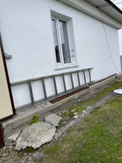 Outside a building in Talmenka, Russia, a gray wooden ladder leans against a white exterior wall. A window is above the ladder, and a brown roof edge is below. A concrete curb runs along the base of the wall, with grass and gravel beyond the curb.