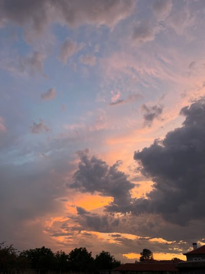The image captures a dramatic sunset over the city of Skutare, Bulgaria. The sky is filled with a vibrant display of colors, ranging from deep oranges and yellows where the sun is setting, to soft pinks and blues higher in the atmosphere. Dark, heavy clouds are interspersed with thinner, wispy formations, creating a dynamic and textured sky.
In the foreground, the silhouettes of lush green trees are visible, their branches reaching upwards towards the colorful sky. Below the trees, the rooftops of houses can be seen, with one house prominently featuring a red-tiled roof and a chimney. The overall scene evokes a sense of natural beauty and the tranquil end of a day in a residential area of Skutare. There are no people or active events depicted in the image.