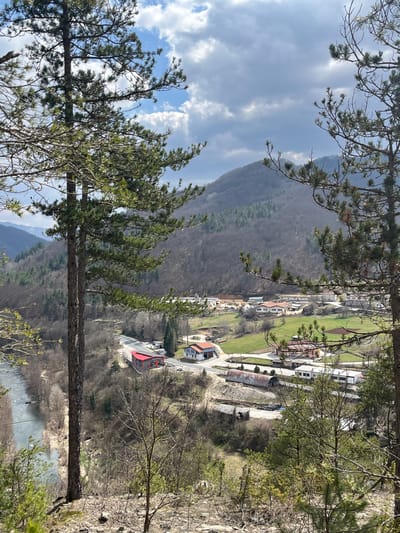 The image presents an elevated panoramic view of a valley in Srednogortsi, Bulgaria. The sky is partly cloudy, displaying both bright blue areas with white cumulus clouds and darker grey cloud formations. In the foreground, several pine trees with dark trunks and green needles, alongside bare deciduous branches, frame the scene from a rocky, sparsely vegetated slope.
A light blue-grey river is visible in the lower-left midground, flowing alongside a paved road. The road curves sharply to the right, revealing a modern-looking building with a prominent red roof and grey walls positioned adjacent to the curve. Further into the midground, a scattering of structures typical of a small settlement is present, including several white-walled houses with orange/red tiled roofs. An industrial area features a long, corrugated metal arch-shaped building and other smaller structures, with evidence of cleared land or construction nearby. Green grassy fields are interspersed between the buildings and industrial zones.
The background is dominated by forested mountains, appearing primarily deciduous with patches of conifers, suggesting a late winter or early spring season due to the bare tree branches on many slopes. No individuals or specific activities are discernible within the scene.