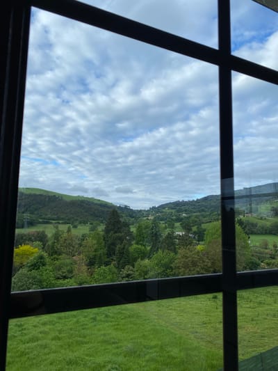 View through a multi-pane window showing a landscape. The landscape includes rolling hills covered in green grass and trees under a sky with scattered white clouds. The location is likely the countryside near Villaviciosa, Spain given the geography and vegetation. No people are visible.