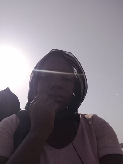 The scene depicts a single individual, appearing female, positioned outdoors in Ibi, Nigeria. The subject is captured from the chest up, silhouetted against a bright, overexposed sky, with strong direct sunlight originating from the upper left, causing significant lens flare across the subject's face. The individual wears a dark, sheer head covering. Facial features are obscured by shadow and glare. A light-colored, possibly pink or light brown, short-sleeved top with a visible texture is worn. The subject's right hand (viewer's left) is raised, with fingers curled and the knuckles resting near the chin or lower mouth area. A dark earbud cable is visible extending downwards from the subject's right side (viewer's left). To the far left, another indistinct, dark object is partially visible, also silhouetted against the bright sky. The environment is characterized by an open, outdoor setting with a clear, bright sky.