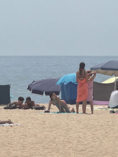 The image depicts a beach scene with several individuals. In the foreground, a woman is positioned on her hands and knees on the sand, wearing a light-colored swimsuit. To her left, two men are lying on the sand, appearing to be looking at a mobile phone. Further to the left, a person's legs are visible lying on a towel. In the mid-ground, two women are standing, one with a dark braid is wearing an orange wrap, and the other, wearing a pink wrap, has her arms raised. Behind them, beach umbrellas are visible, one blue and one dark purple. The background consists of the sea and a hazy sky, suggesting a clear, overcast day. The location is the city of Espinho, Portugal, known for its sandy beaches and popular boardwalk.