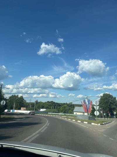 The image shows a sunny day in Vereya, Russia, with a clear blue sky dotted with fluffy white clouds. The perspective is from inside a vehicle, looking out at a road intersection. The road curves to the left, with a light gray car driving away from the camera. On the right side of the intersection, there is a monument with several flags, possibly Russian flags, attached to it. The flags are red, white, and blue. There are also traffic signs and streetlights visible. The area around the intersection is green with trees and grass. In the background, there are some buildings and more trees. The overall impression is a rural or suburban setting on a beautiful summer day.