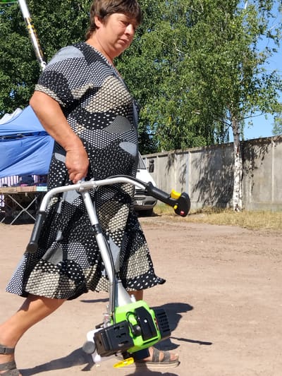 A woman in a black and white patterned dress is walking with a green string trimmer in the city of Irshansk, Ukraine. She is holding the trimmer by its handle. The trimmer has a silver shaft and black handles. The woman is wearing sandals. In the background, there is a blue tent, a table with items on it, a white van, a concrete fence, and trees. The ground is dirt. The sky is blue.