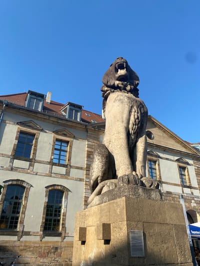 A low-angle, color photograph shows a large stone lion statue and sections of two buildings. The lion, positioned in the upper-center, is roaring with an open mouth and visible teeth. It is situated on a stone plinth. To the right and behind the statue, parts of a building with gray facade and framed windows are visible. A building with a red tile roof and dormer windows is partially visible in the left background. The sky is a bright, clear blue. There is some writing on the stone plinth beneath the statue. This is the Löwendenkmal (lion monument) in Landau in der Pfalz, Germany.