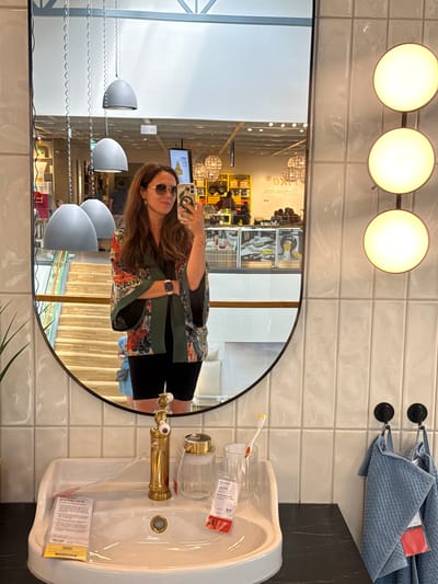 This is a portrait-oriented, slightly angled, full-body selfie of a woman standing in a modern bathroom or showroom. The woman is in her late 20s or early 30s, with long brown hair, wearing sunglasses and a dark floral print kimono-style top over a black outfit. She is holding a smartphone in her right hand and appears to be taking the photo using the phone's front-facing camera.
The setting features a large, oval mirror that reflects the woman and the surrounding environment. Behind her, the wall is tiled with a light-colored, glossy subway tile. Above the mirror, a modern light fixture with three circular, illuminated globes is visible. To the right of the mirror, a white sink with a gold-colored faucet is mounted on a dark countertop. On the sink, there is a clear cup with a toothbrush and another clear container, likely for storage. Several white tags with red text are attached to the objects. Above the sink, a stack of folded blue towels is hanging from black hooks on the tiled wall.
The reflection in the mirror offers a view of the space behind the woman. It shows a brightly lit interior with shelves stocked with various items, suggesting a retail environment such as a home goods store or a showroom. The reflections of lighting fixtures and displays are prominent.
The image appears to be taken indoors during the daytime, with ample artificial lighting. The mood seems casual and relaxed. Text visible on the tags includes "PUTSION" and "IKEA". The location context is Ulbroka, Latvia.