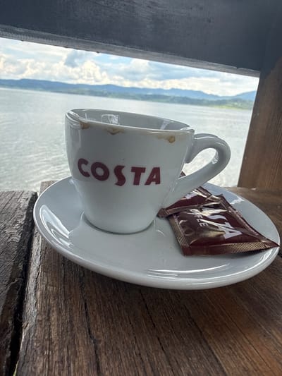 A white ceramic Costa coffee cup, stained with coffee residue around its rim, sits on a matching white saucer. Two dark red sachets, branded "COSTA COFFEE" and likely containing sugar or sweetener, rest on the saucer beside the cup. This ensemble is positioned on a rustic, weathered dark brown wooden surface, possibly a table or railing, displaying visible grain patterns and cracks. In the background, a serene expanse of calm, grey-blue water, identifiable as Batak Lake, stretches towards the horizon. Distant, hazy blue-green mountains and hills line the far shore of the lake. The sky above is partially cloudy, featuring white cumulus clouds against a pale blue backdrop. The scene is framed at the top and right by dark wooden beams, suggesting an outdoor viewing or seating area overlooking the lake in Цигов чарк, Bulgaria. No individuals are present in the image.