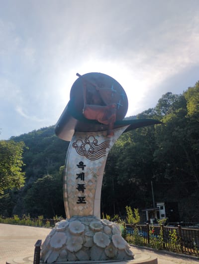 A tall, ornate monument stands prominently in the center of the frame, set against a backdrop of densely forested mountains under a bright, partly cloudy sky. Strong sunlight emanates from the upper right, causing a lens flare and backlighting the top of the structure.

The monument's base is adorned with multiple light-colored, stylized floral or leaf-like shapes. Rising from this base is a tall, silver-toned column featuring a textured, possibly woven or checkered, pattern. On this column, a large, stylized brown cloud motif is visible, below which are prominent black Korean characters reading "옥계폭포" (Okgye-pokpo), identifying the site as Okgye Waterfall. The upper section of the monument comprises a dark, circular disk with several light blue cross-shaped symbols, supporting a dark, stylized figure that appears to be holding a staff or instrument.

The monument rests on a paved pathway or plaza. To the left, a portion of a tree with green leaves is visible. The background is dominated by steep, lush green mountains covered in dense foliage. To the right of the monument, a low, dark wooden fence runs parallel to the path, with scattered green plants and a few red flowers behind it. Further in the background on the right, a small, white building with a brown roof and a porch is partially visible amidst the trees. No people are present in the scene, which is located in the vicinity of Godang-ri, South Korea.