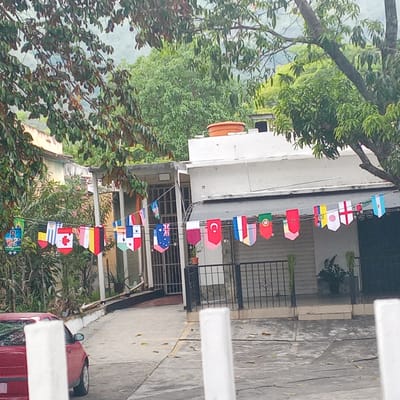 The image captures an outdoor scene with a residential building featuring a white facade and a white roof, partially obscured by lush green trees. A string of international flags decorates the front of the building, strung between poles and the structure itself. The flags appear to represent various countries. In the foreground, a red car is parked on a paved surface. The environment suggests a warm climate, with foliage and what looks like a slightly overcast sky. The location is identified as El Limón, Venezuela.