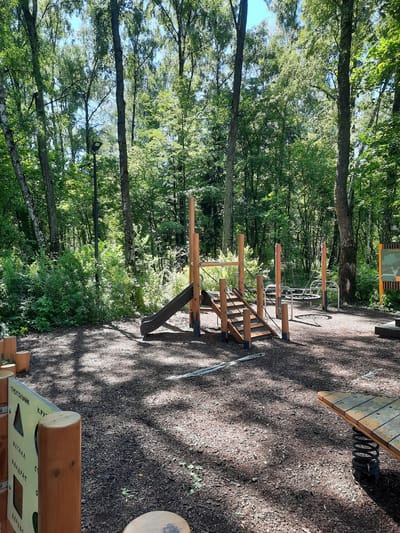 The image depicts a modern, natural-themed playground situated in a forest environment. The playground features wooden structures including a slide, stairs, climbing frames, and a circular rope swing. Educational elements are also present, such as a panel with shapes like a triangle, circle, and square, labeled in Russian. The ground is covered with wood chips, and there are scattered wooden log seats and a spring-loaded wooden rideable toy. The surrounding environment is a dense forest with tall trees, dappled sunlight filtering through the leaves, and a blue sky visible in the background. In the distance, a lamppost is visible amongst the trees. The playground appears to be located in Mar'ino, Russia, a district within the southeastern administrative okrug of Moscow, known for its green spaces and parks. No people are present in the image.