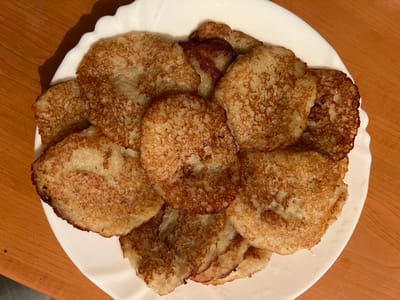 A top-down, medium shot captures a white, scalloped-edged plate filled with numerous golden-brown potato pancakes, known as bramboráky in Czechia. The pancakes are unevenly shaped and stacked atop one another, with some appearing slightly crispier than others, indicating they have been recently fried. The plate rests on a warm-toned wooden surface, likely a table. The lighting suggests an indoor setting, possibly a kitchen or dining room. The image is devoid of any people or distinct architectural features that would pinpoint a specific location within the city of Náchod, Czechia. The focus is solely on the presentation of the food.