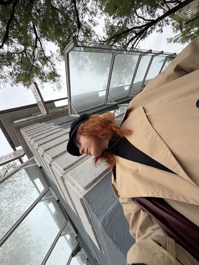 The image is a low-angle shot of a person with red hair wearing a black hat and a beige trench coat. The person is positioned in front of a building with multiple glass windows. The windows appear wet. In the background, trees with green leaves can be seen. The photo was taken in Kehl, Germany, indicated by the "dm" sign, a German drugstore chain.