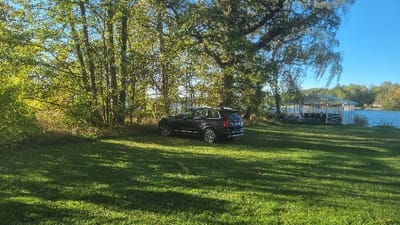 The image presents a static outdoor scene on a clear, sunny day, located at the waterfront area near Sjökrogen restaurant at Gränsö Slott in Västervik, Sweden.
In the mid-ground, a dark-colored Volvo XC90 SUV is parked stationary on a well-maintained green lawn. The vehicle is positioned with its rear-left side visible, showcasing multi-spoke alloy wheels and roof rails. Behind the vehicle and to the left, a dense cluster of deciduous trees with green foliage forms a natural backdrop, partially obscuring the water beyond. A large, mature tree with a spreading canopy is prominent in the upper right quadrant of the image.
A calm body of water, consistent with a bay or inlet, is visible extending across the mid-ground behind the trees and prominently on the right side. Directly situated over this water body, on the right side of the frame, is a white, multi-sided gazebo-like structure. This structure features a white roof supported by white pillars and is enclosed by white railings. Inside the gazebo, dark-colored tables and chairs are arranged.
In the distant background, across the water, additional green foliage is visible along with a small white building featuring a distinct red roof, likely a house or cabin. The sky is clear and bright blue. Strong, direct sunlight illuminates the scene, casting pronounced shadows from the trees and the parked vehicle onto the grass in the foreground. No individuals are present in the image.