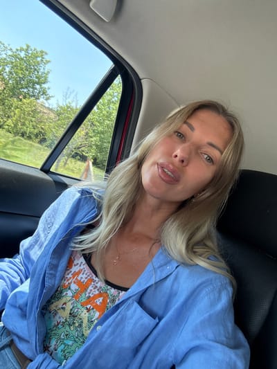A woman with blonde hair is taking a selfie inside a vehicle, with the window open to a rural scene. She is wearing a light blue unbuttoned shirt over a colorful patterned top and a delicate necklace. Her hair is partially obscuring her face as she looks towards the camera. The background through the open window shows green trees and a bright blue sky, suggesting it is daytime with clear weather. The interior of the car is visible, including the headrest and part of the seat. The scene suggests a casual outing or travel. There is no visible text in the image. The location context provided is Radanovići, Montenegro.