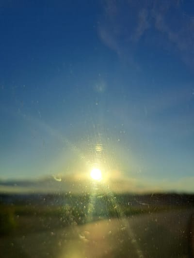 The image shows a bright sun low in the sky, casting a warm glow. The sun appears to be shining through a dirty or streaked window, creating lens flares and distorting the view of the landscape below. The landscape itself is out of focus, appearing as a blurred band of greens and browns, suggesting it is likely a natural or rural setting rather than an urban one. The sky is a clear blue, with a few wispy clouds. There are no people, animals, or specific man-made structures clearly visible. Given the lack of discernible urban elements, it is not possible to determine if this scene is located in the city of Chernetsk, Russia. The image composition and focus suggest it might be a view from a vehicle or a window in a rural or suburban area.