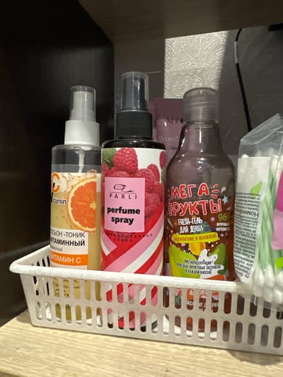 The image displays a white plastic basket containing various cosmetic products, likely stored on a shelf. Prominently featured are three bottles with spray or pump dispensers. The leftmost bottle is transparent and contains a yellowish liquid, with a label showing an orange slice and the text "Vitamin C" in English and Russian. To its right is a pink bottle with a label featuring raspberries and the text "parli perfume spray" in English and Russian. The third bottle, to the right of the perfume spray, is transparent and filled with a brown liquid, featuring a label with colorful graphics and Russian text including "MEGA FRUITS" and "FRESH-GEL FOR SHOWER". Behind these bottles, partially visible, are other items, including a pink tube and a bag of cotton swabs. The background consists of dark shelving. The basket rests on a light-colored wooden surface. The setting appears to be domestic or a small retail environment. There are no people or animals present in the image. The city of Shaumyan, Russia, is not depicted in the image.