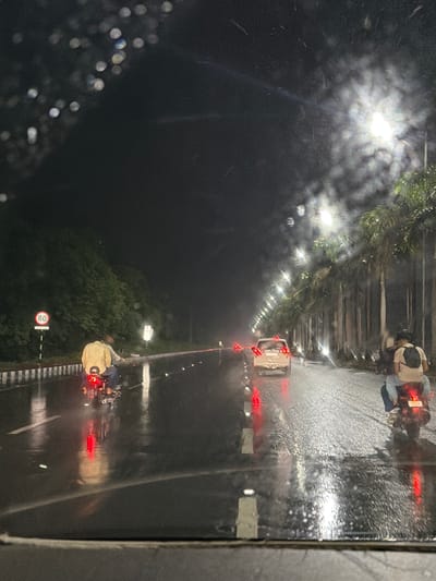 The image depicts a nighttime scene on a wet road in Gollapalle Khurd, India, likely during or shortly after a rain shower, given the reflections on the asphalt and visible water droplets on what appears to be a windshield. The road is lined with palm trees on the right side, illuminated by streetlights. Two motorcycles are visible on the road. The first motorcycle, in the foreground and left of the frame, is ridden by a person wearing a light-colored jacket and blue jeans. The motorcycle's taillight is illuminated red. A circular traffic sign indicating "80" is present on the left side of the road. Further down the road, another motorcycle is visible with two occupants, the rider and a passenger wearing a backpack. A white car is also on the road, ahead of the second motorcycle. The overall atmosphere is dark, with the bright streetlights creating reflections and a somewhat hazy effect on the wet surface.